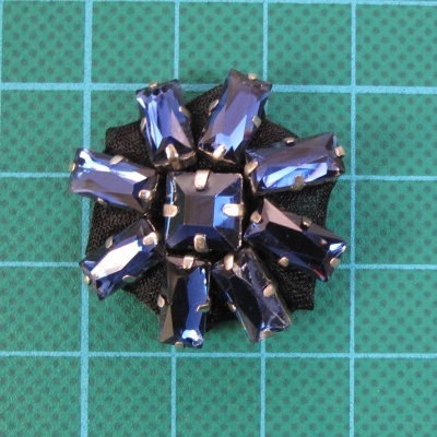 Broche flor com cristais azuis em base preta sobre superfície quadriculada verde