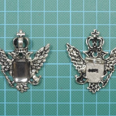 Emblemas metálicos prateados com asas e coroas sobre superfície verde quadriculada