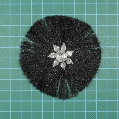 Acessório circular com cerdas pretas e flor de pedras transparentes no centro sobre fundo azul em malha de corte