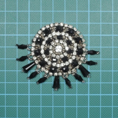 Broche circular com pedras prateadas e esferas pretas sobre superfície quadriculada verde