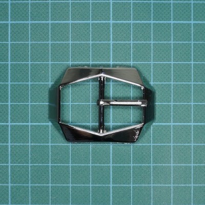 Fivela metálica prateada com design geométrico sobre fundo verde quadriculado