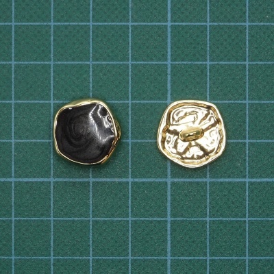 Dois botões quadrados dourados, um preto na frente, outro dourado atrás, sobre superfície verde com grelha.