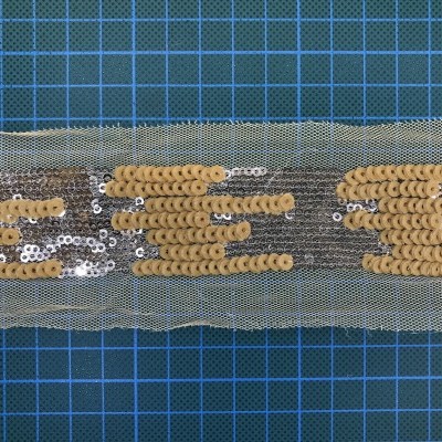 Fita decorativa em rede bege com lantejoulas douradas e prateadas sobre fundo azul quadriculado