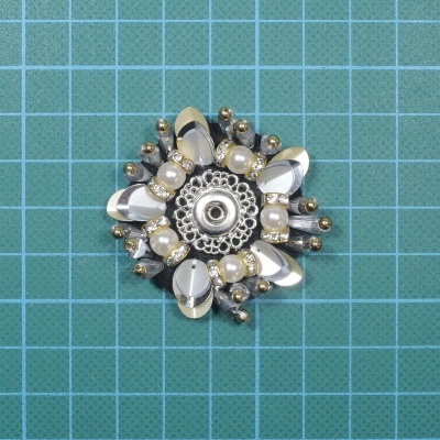 Broche em forma de flor com contas prateadas e pérolas sobre fundo quadriculado verde