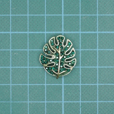 Pendente em forma de folha verde com metal dourado sobre fundo quadriculado azul esverdeado