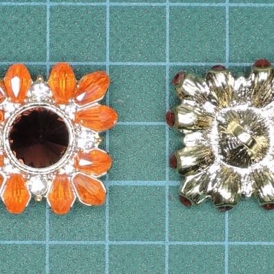 Dois botões metálicos em forma de flor, um laranja e outro dourado, sobre fundo verde quadriculado.