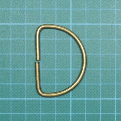 Argola metálica em forma de D dourada sobre base quadriculada verde.