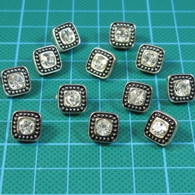 Doze botões quadrados em metal com pedra central sobre fundo verde quadriculado