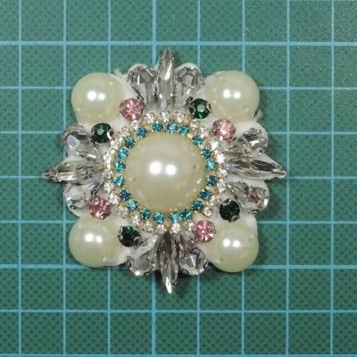 broche com pérolas, pedras coloridas e cristais sobre fundo verde com grelha branca