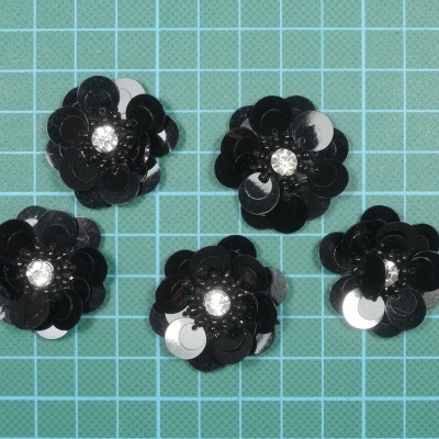 Botões decorativos em formato de flor com lantejoulas pretas e centro brilhante sobre fundo quadriculado