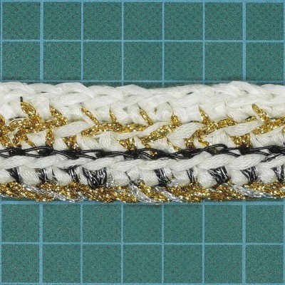 Três fitas decorativas trançadas branca, dourada e preta sobre fundo quadriculado verde