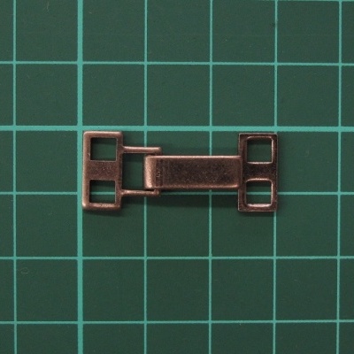 Peça metálica cobreada com aberturas quadradas numa superfície quadriculada verde