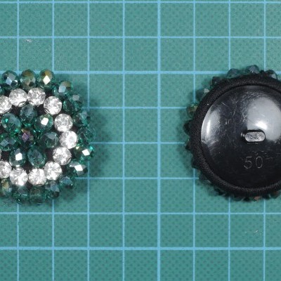 Dois botões decorativos, um com pedras verdes e brancas, outro preto liso sobre superfície verde com grade branca.