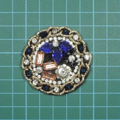 Broche redondo decorativo com pedras azuis, laranjas e brancas sobre fundo azul quadriculado