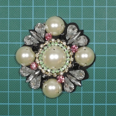 Broche decorativo com pérolas e pedras coloridas sobre fundo quadriculado verde