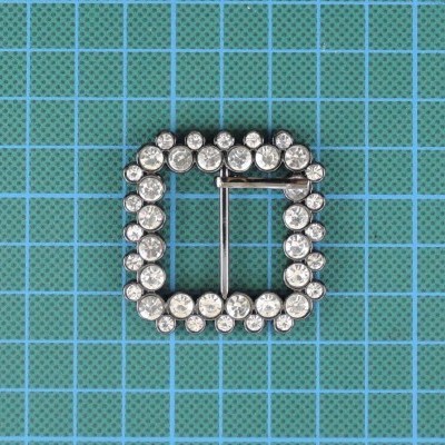 Fivela quadrada prateada com pedras transparentes em fundo azul quadriculado