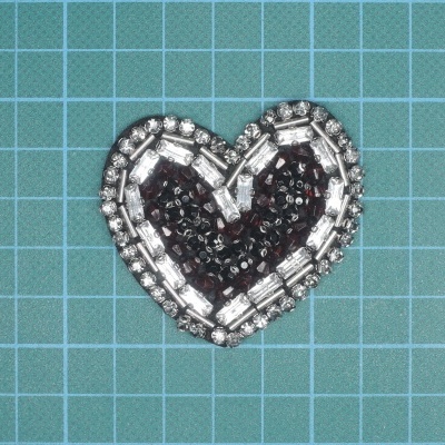 Aplicação em forma de coração com contas pretas e pérolas transparentes em fundo quadriculado azul claro