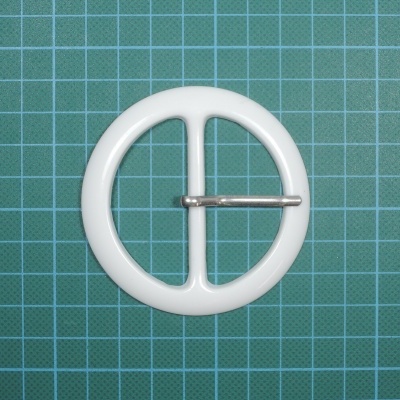 Fivela redonda branca com barra central e pino metálico sobre superfície verde quadriculada.