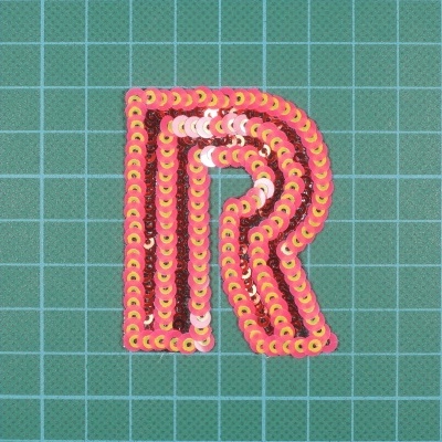 Patch em forma da letra R com lantejoulas rosa e castanho, sobre fundo quadriculado verde