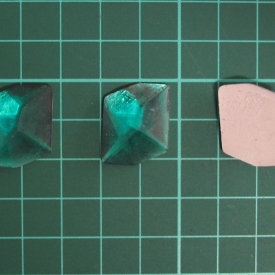 Três peças pequenas sobre base verde quadriculada, duas verdes translúcidas piramidais e uma rosa opaca irregular