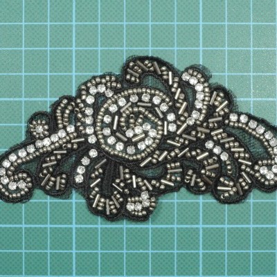 Aplicação decorativa com contas pretas e prateadas em forma floral sobre fundo quadriculado verde