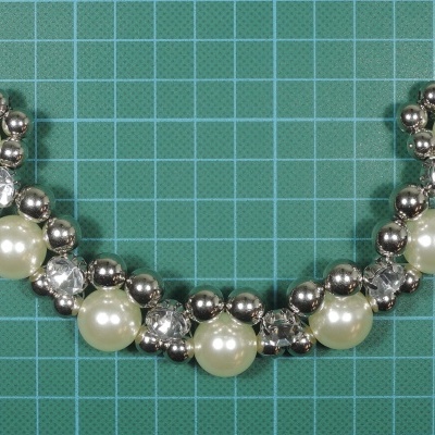 Adereço decorativo com esferas pérola, esferas prateadas e cristais em padrão simétrico sobre fundo verde quadriculado