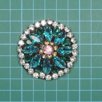 broche redondo com pedras azuis e rosas em moldura prateada sobre superfície quadriculada verde