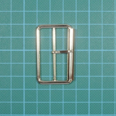 Fivela metálica dourada rectangular sobre superfície quadriculada verde.