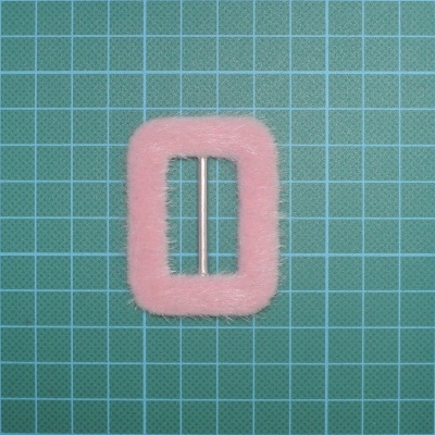 Fivela retangular cor-de-rosa felpuda numa superfície verde com padrão quadriculado.