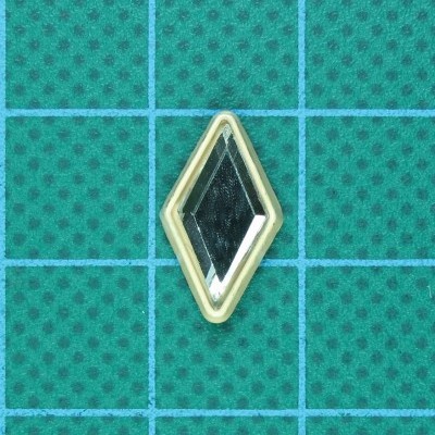 Peça metálica em forma de diamante dourada e preta sobre superfície verde com grelha azul