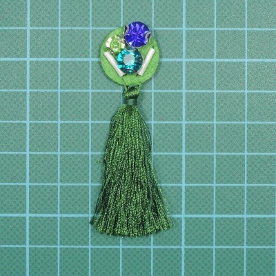 Pingente verde com franjas e pedras brilhantes em fundo quadriculado azul