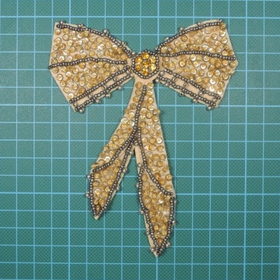 Aplicação de laço decorativo bege com pérolas douradas e contas escuras sobre fundo com padrão de grades