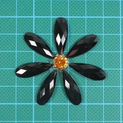 Flor decorativa com pétalas pretas facetadas e centro laranja sobre base quadriculada verde