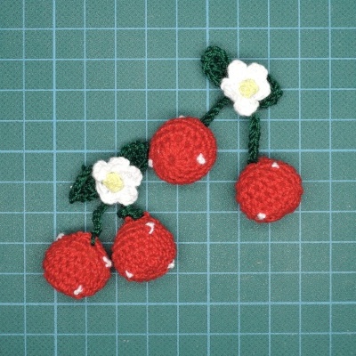 Cerejas vermelhas de crochê com flores brancas e folhas verdes sobre superfície quadriculada azul