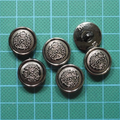 Seis botões metálicos prateados decorados com emblemas circulares sobre papel quadriculado verde