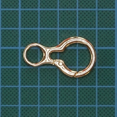 Argola metálica dourada tipo mosquetão sobre superfície verde com grelha