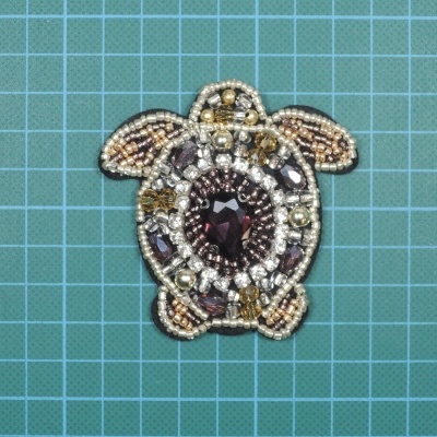 Broche em forma de tartaruga com pérolas e pedras coloridas sobre superfície verde com grelha azul
