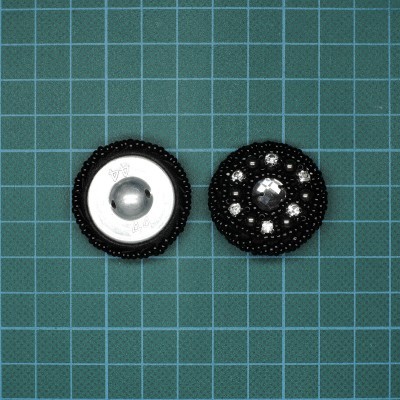 Botões redondos, um metálico prateado e outro preto com pedras brilhantes, sobre superfície quadriculada