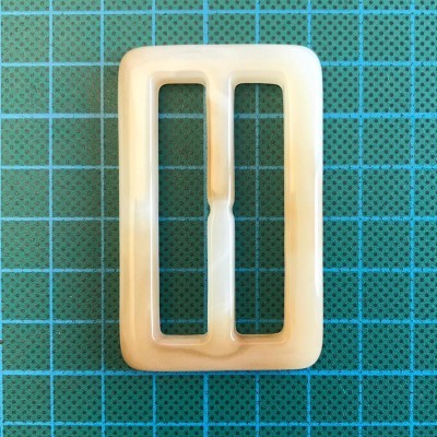 Fivela retangular bege perolado sobre superfície verde com grade azul.