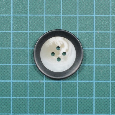 Botão redondo branco com borda preta e quatro furos sobre superfície verde quadriculada