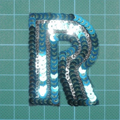 Aplicação de tecido letra R em lantejoulas prateadas e azuis sobre fundo quadriculado verde