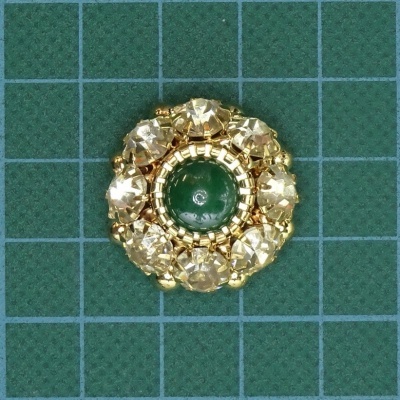 Botão decorativo redondo com pedras douradas e centro verde sobre tecido verde quadriculado
