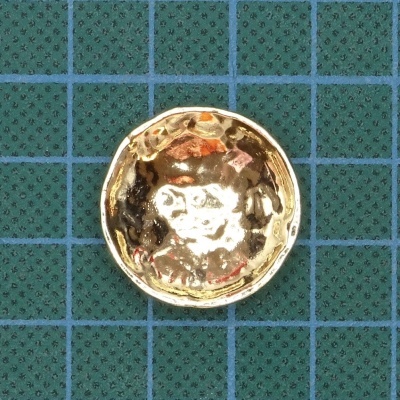 Botão metálico dourado com textura martelada sobre tecido verde com grelha azul