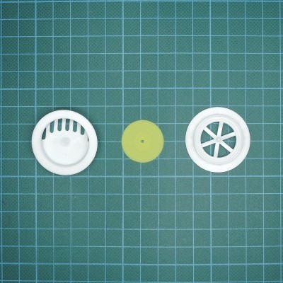 Três peças circulares, duas brancas em plástico e uma verde, sobre superfície verde quadriculada