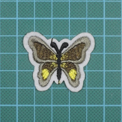 Patch bordado de borboleta com cores castanho, amarelo, preto e branco num fundo quadriculado verde