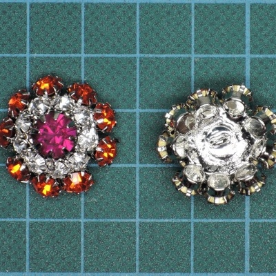 Dois botões decorativos redondos com cristais coloridos e metálicos sobre superfície quadriculada verde