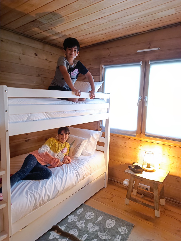 Gift Voucher - Fun Glamping Natura | 2 Noites Beliche branco em quarto de madeira com duas crianças, criado-mudo com candeeiro e tapete cinza com padrão de folhas.