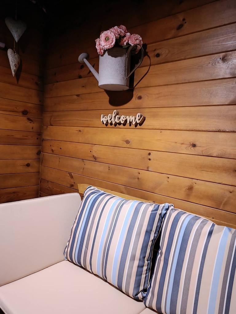Gift Voucher - Fun Glamping Natura | 2 Noites Sofá branco com almofadas às riscas azuis junto a parede de madeira com regador com flores e placa 'welcome'