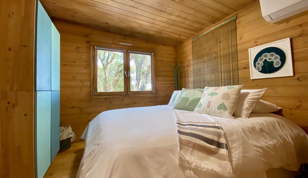 Gift Voucher - Fun Glamping Natura | 2 Noites Quarto com cama de madeira, roupa branca e almofadas verdes, paredes e teto de madeira, janela com vista para árvores