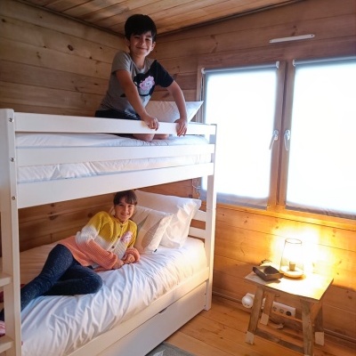 Beliche branco em quarto de madeira com duas crianças, criado-mudo com candeeiro e tapete cinza com padrão de folhas.